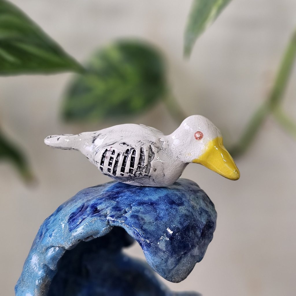 A close up shot of a ceramic sculpture of a bird atop a wave. The bird is white, with black vertical stripes carved into its wing. It has a reddish brown eye, and a big yellow beak. Its feet are tucked away, and its tail sits horizontally.