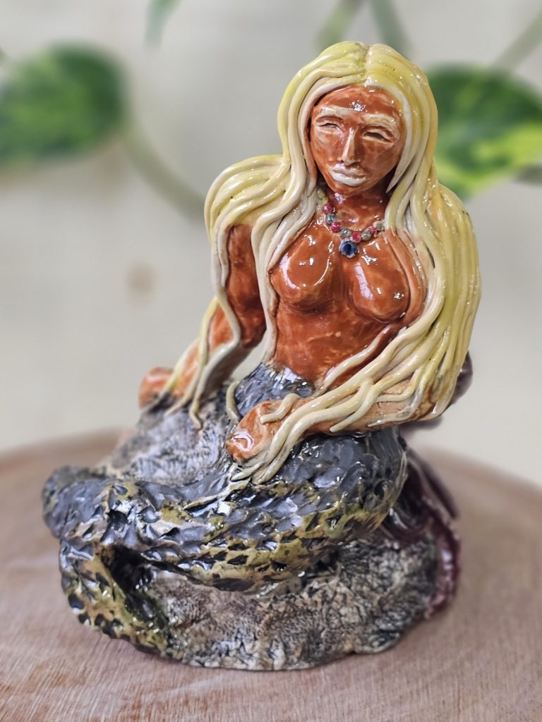 A sculpture of a mermaid sitting atop a rock, one hand holding the rock and the other across her lap, with her tail curving down and round the side of the rock. The mermaid has thick strands of yellow hair, which flow over her shoulders, down her arms and over her hands. She has full lips and wears a calm expression. She wears a beaded necklace of green, red and blue, and no top. Her tail is green and black, with an intricate carved texture of scales. The rock is dappled grey, jagged in placed and smoother in others.