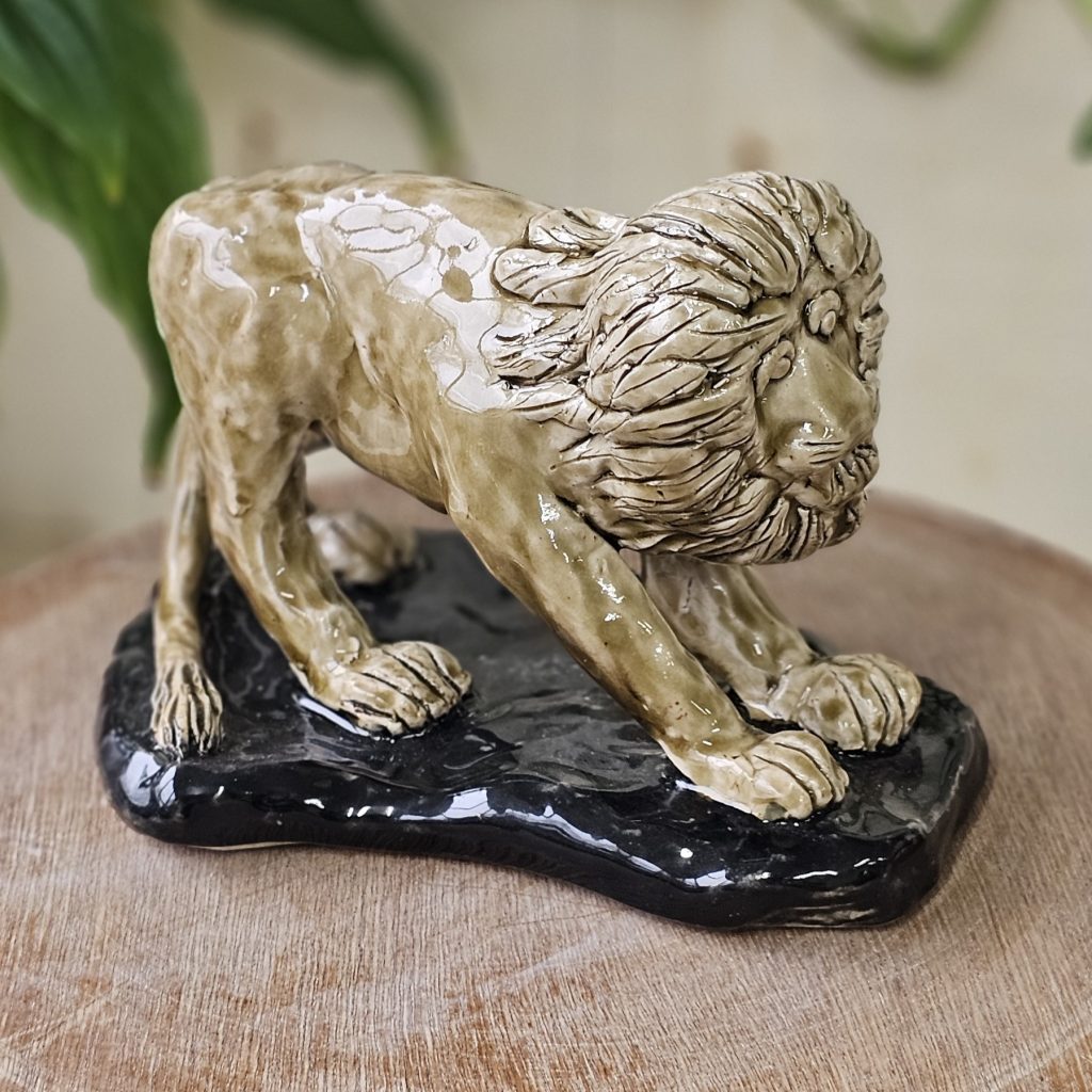 A sculpture of a lion upon black ground. The lion is a pale gold. The lion leans back, so that his weight is more on his back legs, with the front legs stretched out. The lion has a magnificent textured mane and open, curious eyes.