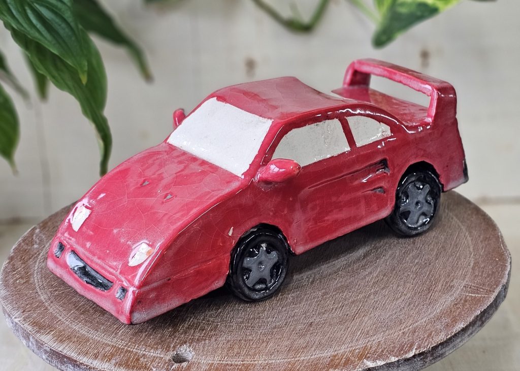 A ceramic sculpture of a red racing car. The car is sleek and streamlined, with a spoiler at the back