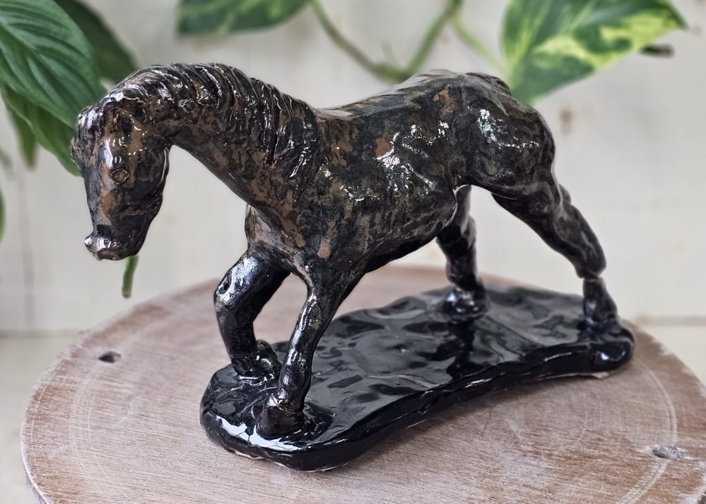 Aceramic sculpture of a horse. It is a mottled, lustrous black brown colour. Its front right leg is raised, and its back legs are outstretched, as though it is setting off to run