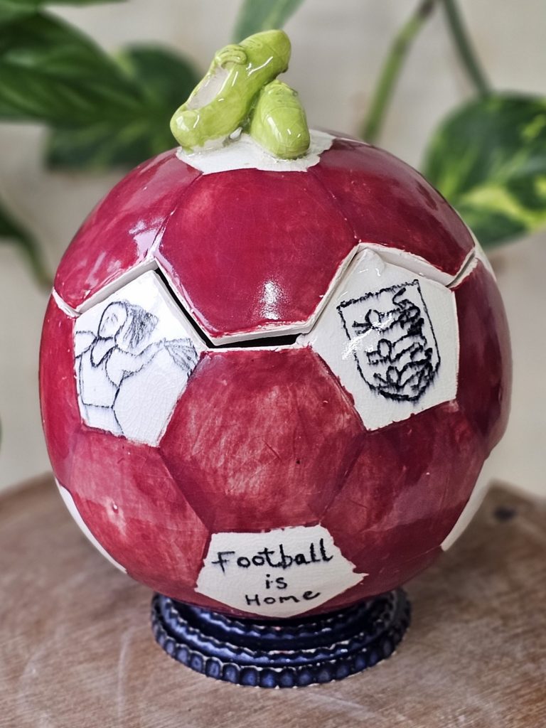 A ceramic vase in the shape of a football. The vase lid is cut along the lines of the red hexagons and white pentagons which make up the football. One pentagon has an image of the three lions on it, another has 'football is home' on it, and another has a picture of one of the England women's football team, or 'lionesses', on it. On top of the football is a small pair of football boots, which act as the handle for the lid. The vase sits on a ridged blue base.