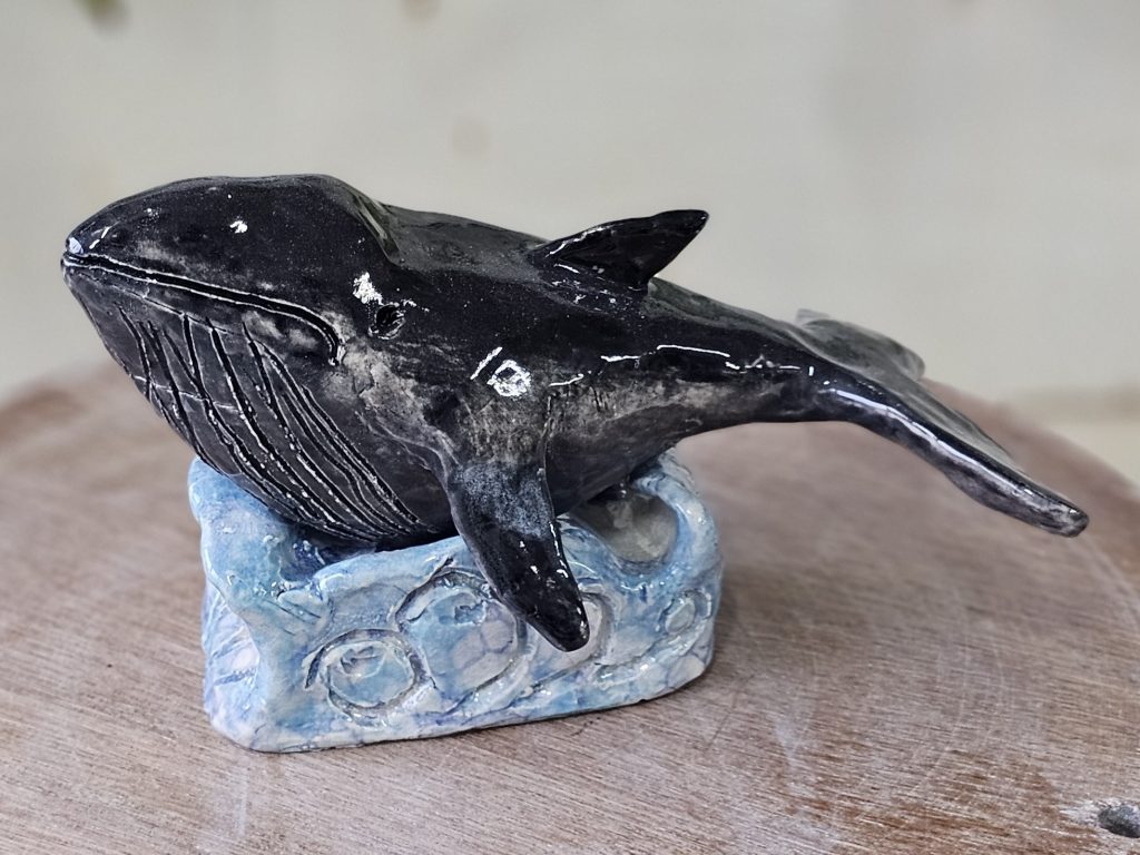 A ceramic sculpture of a minke whale. The Whale is resting atop a plinth sculpted to look like waves. The whale is variously black and dark gray, and has lines of ridges under its mouth along its neck and upper belly. The whale's tail is down and its head is up, as though rising in the water.