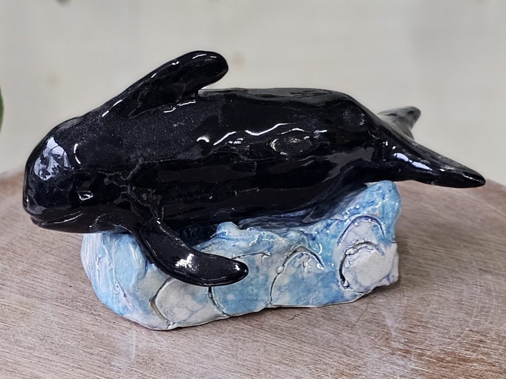 A ceramic sculpture of a pilot whale. The Whale is resting atop a plinth sculpted to look like waves. It is black and has a small curved dorsal fin. The whale looks peaceful, as though it might be resting.