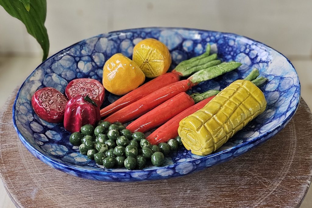 A ceramic sculpture of a plate of food. There are vibrant orange carrots, small round green peas, a lemon cut in half, and a corn on the cob. The plate has high sides and is glazed using a bubble glazing technique, which has created an irregular pattern of circles in various shades of blue. It looks good enough to eat!