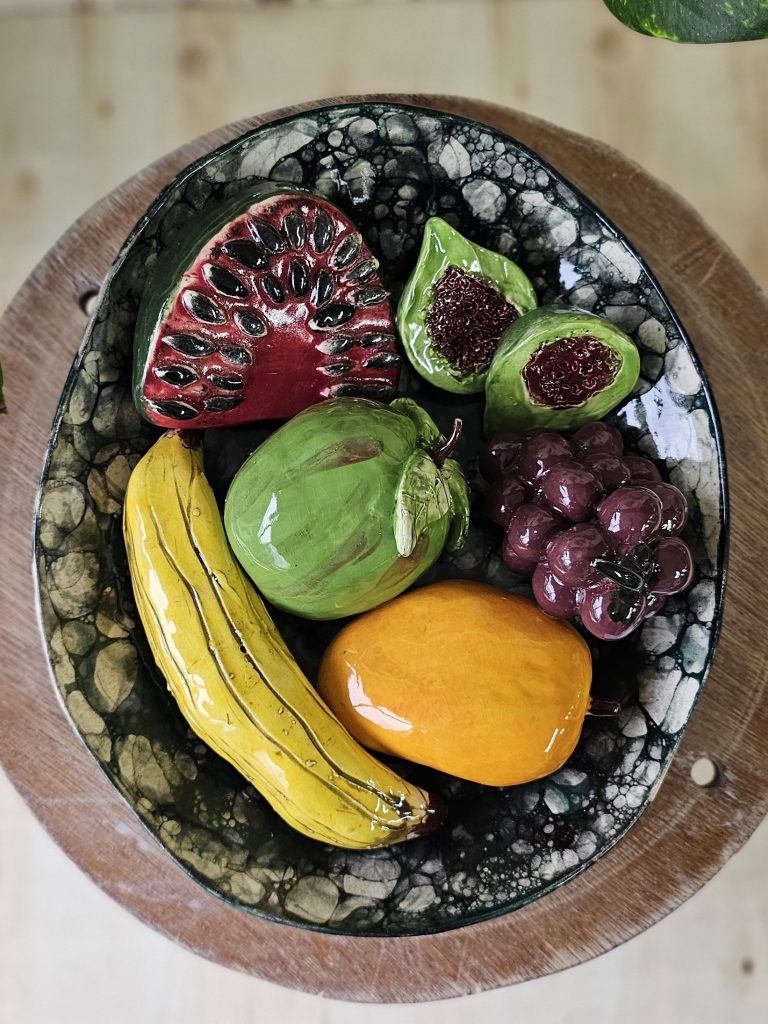 A Ceramic sculpture of a bowl of fruit. There is a slice of watermelon, a banana, a bright orange mango, a green apple with a red blush, a bunch of red grapes, and a fig, cut in half. The bowl has high sides and is glazed using a bubble glazing technique, which has created an irregular pattern of circles in various shades of black, white and grey.