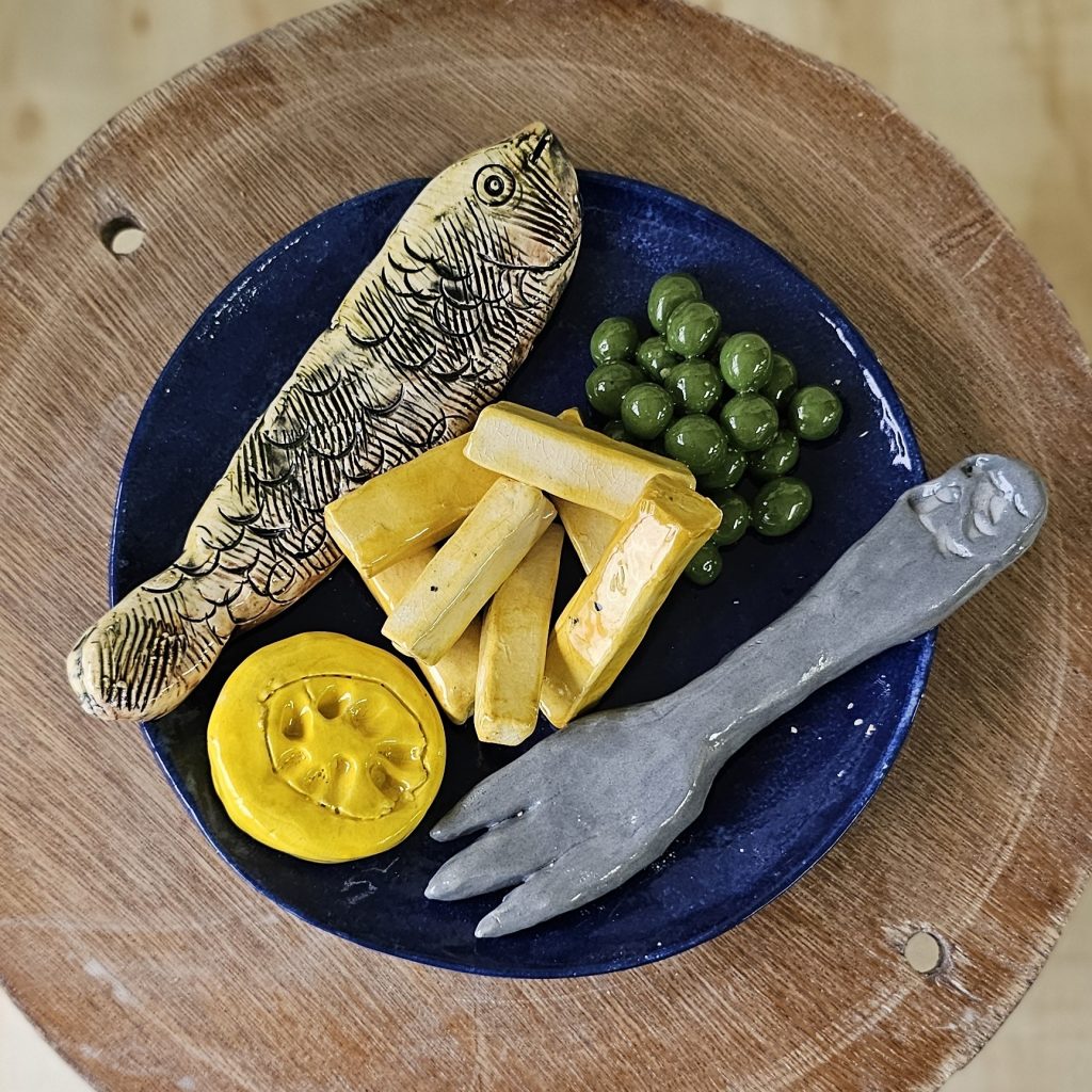 A ceramic sculpture of a plate of fish and chips. The fish is whole, and its surface is textured with the curves and ridges of scales. The chips ore thick cut and stacked in the centre of the plate. There is half a lemon, waiting to be squeezed, a small pile of green peas, and a fork ready to eat them with. The plate is a deep blue.