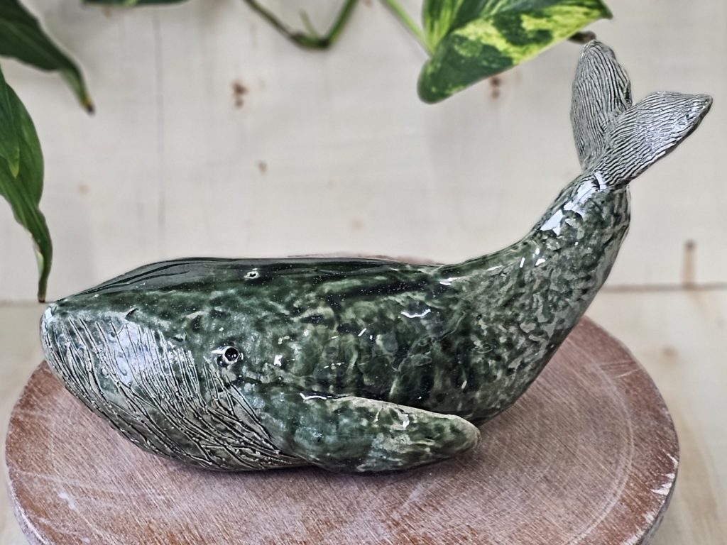 A dark green ceramic sculpture of a whale. The whale has a high wide mouth, with ridges underneath which run along the underside of its body. It has a thin low on either side of its body. and a large tail which is also striped with ridges. The tail is lifted high, as if it might slap down and create a big splash of water.