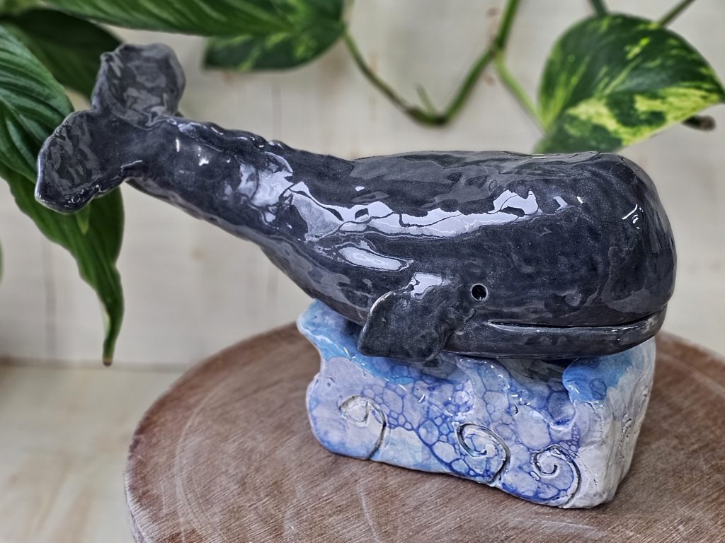 A ceramic sculpture of a sperm whale, sat atop a ceramic plinth sculpted to look like waves. The whale is grey, and has a large square head. Its wide mouth looks almost as though it is smiling. It has a small fin wither side of its body, and a wide tail which is curved slightly upwards as though in the motion of swimming.