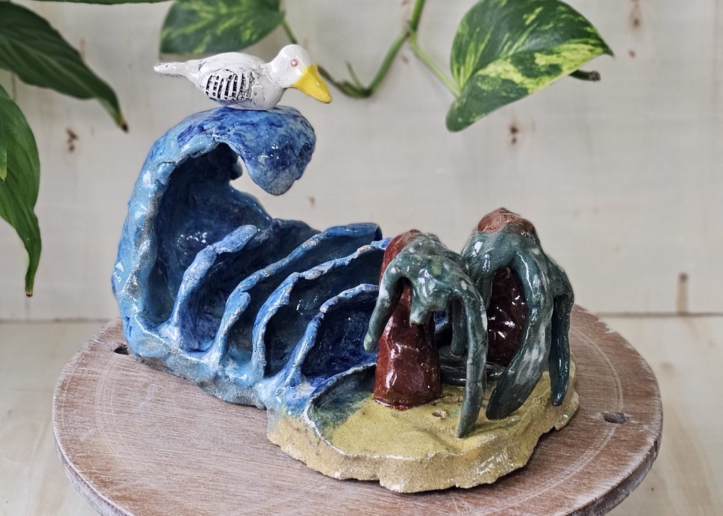A sculpture of waves crashing onto a beach. The waves are a mottled sea blue, and a large wave curls over four smaller ones as they approach the beach. On the shore of the sandy beach, two palm trees droop over, the ends of their leaves touching the sand. Atop the largest waves sits a calm white bird, with black detailing on the wings and a large yellow beak.