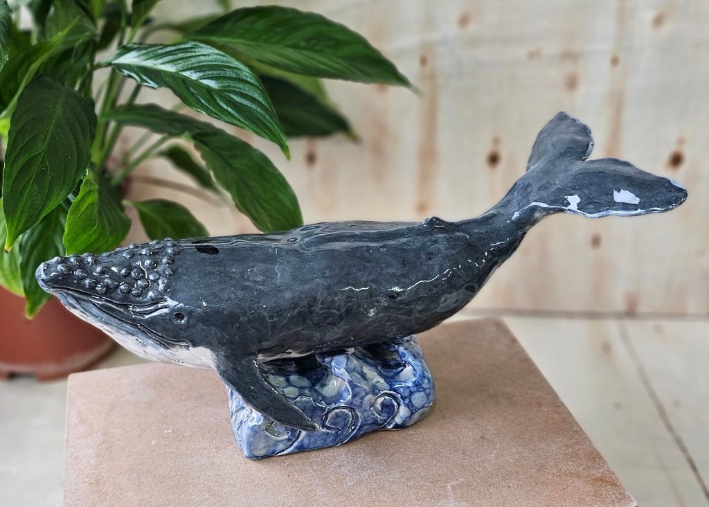 A ceramic sculpture of a hump back whale, sat atop a ceramic plinth shaped as waves. The whale is grey with a white underbelly, and has bumpy ridges on top of its head. Its tail curves upwards, giving the impression that it is in the motion of swimming