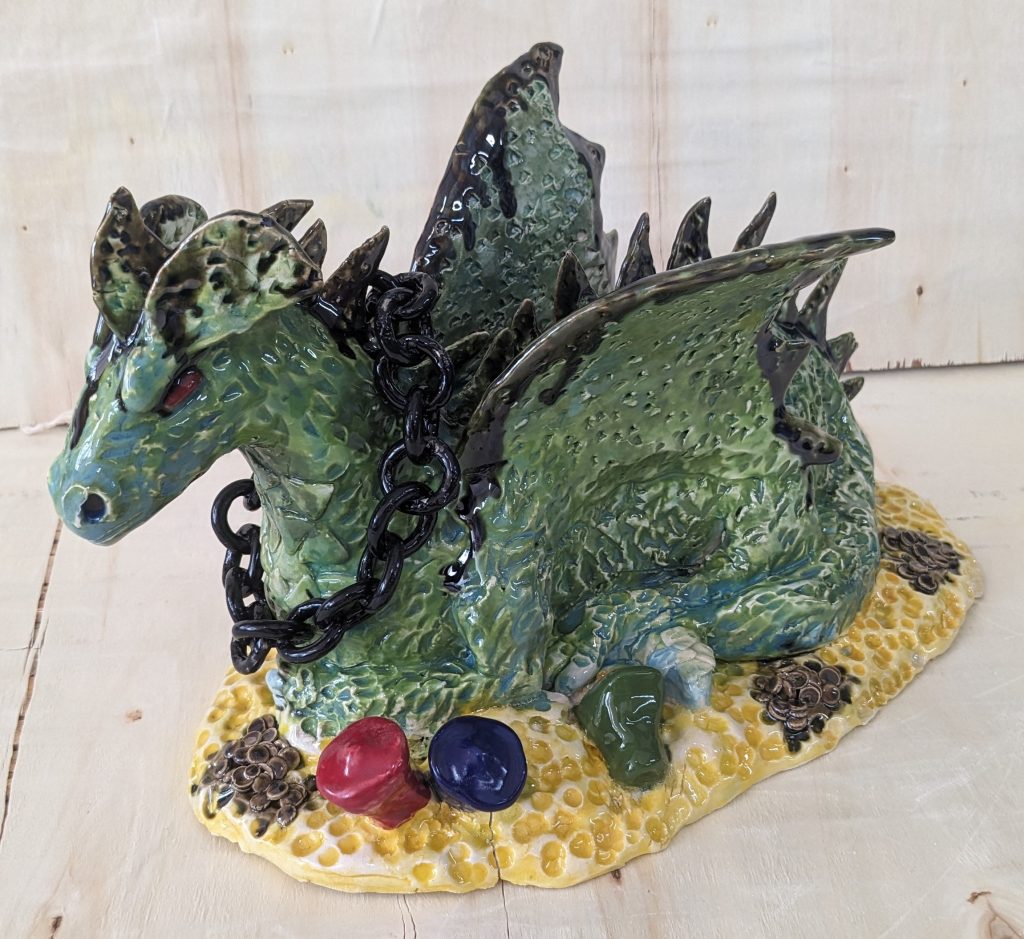 A ceramic sculpture of a dragon, which is dark green with black detailing on its wings, ears, and the spines along its back. The dragon has a chain around its neck, and is sat atop a pile of gold coins and gemstones.