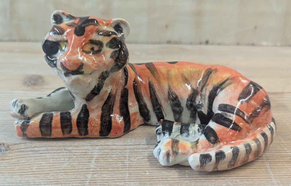 A ceramic sculpture of a tiger, sat with its front paws crossed in front of it, and its back legs curved round its body. It looks curious but relaxed. It is orange and white with black stripes and yellow eyes.