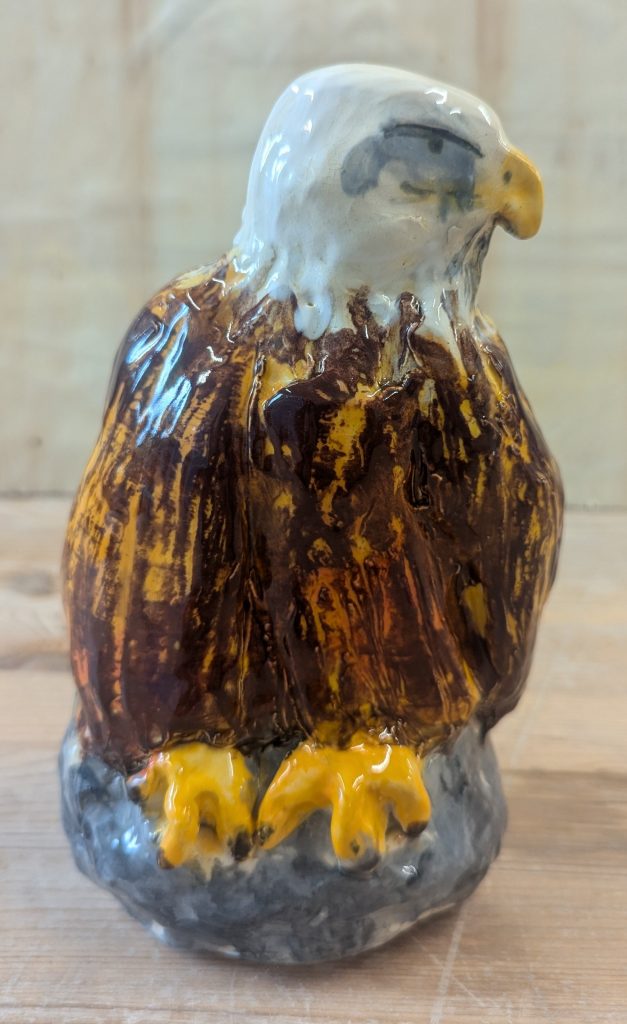 A ceramic sculpture of a golden eagle, perched on a rock, wings to its sides. The eagle is looking to the right, and has a severe expression. Its head is white, its wings a textured golden brown, and its talons are yellow.