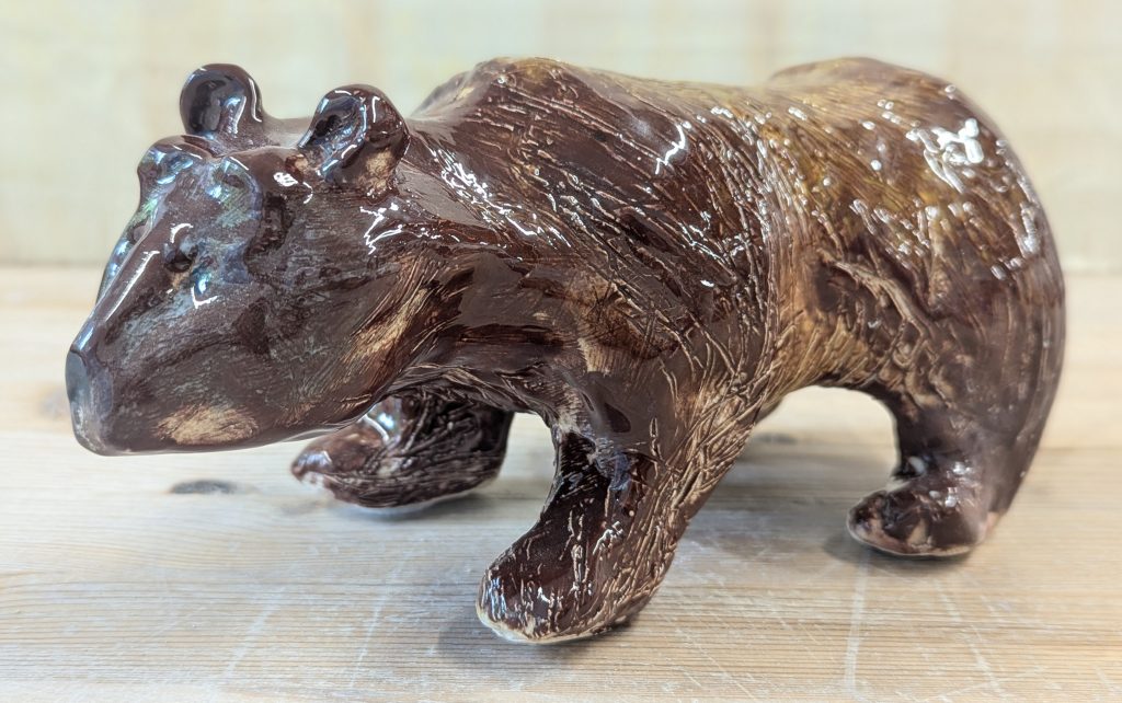 A ceramic sculpture of a brown bear. The bear is standing on all fours. It has small ears, expressive eyebrows and a rounded nose. It is mottled shades of brown, with scratches into the clay creating the texture of fur.