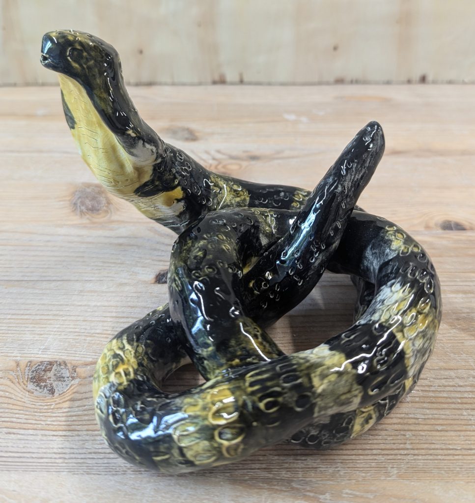 A ceramic sculpture of a snake with its head reared and its body coiled around itself in a knot. The snake is stripey black and yellow and ridged scales along its body.