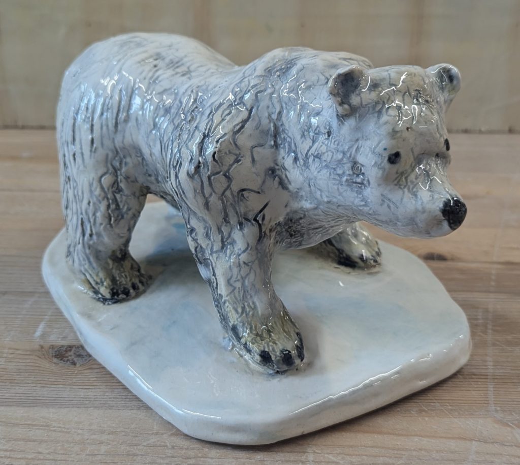 A ceramic sculpture of a polar bear standing on a base of icy snow. The bear has textured fur with grey-blue shadows. It has an inquisitive expression, as though it has paused to look at something.